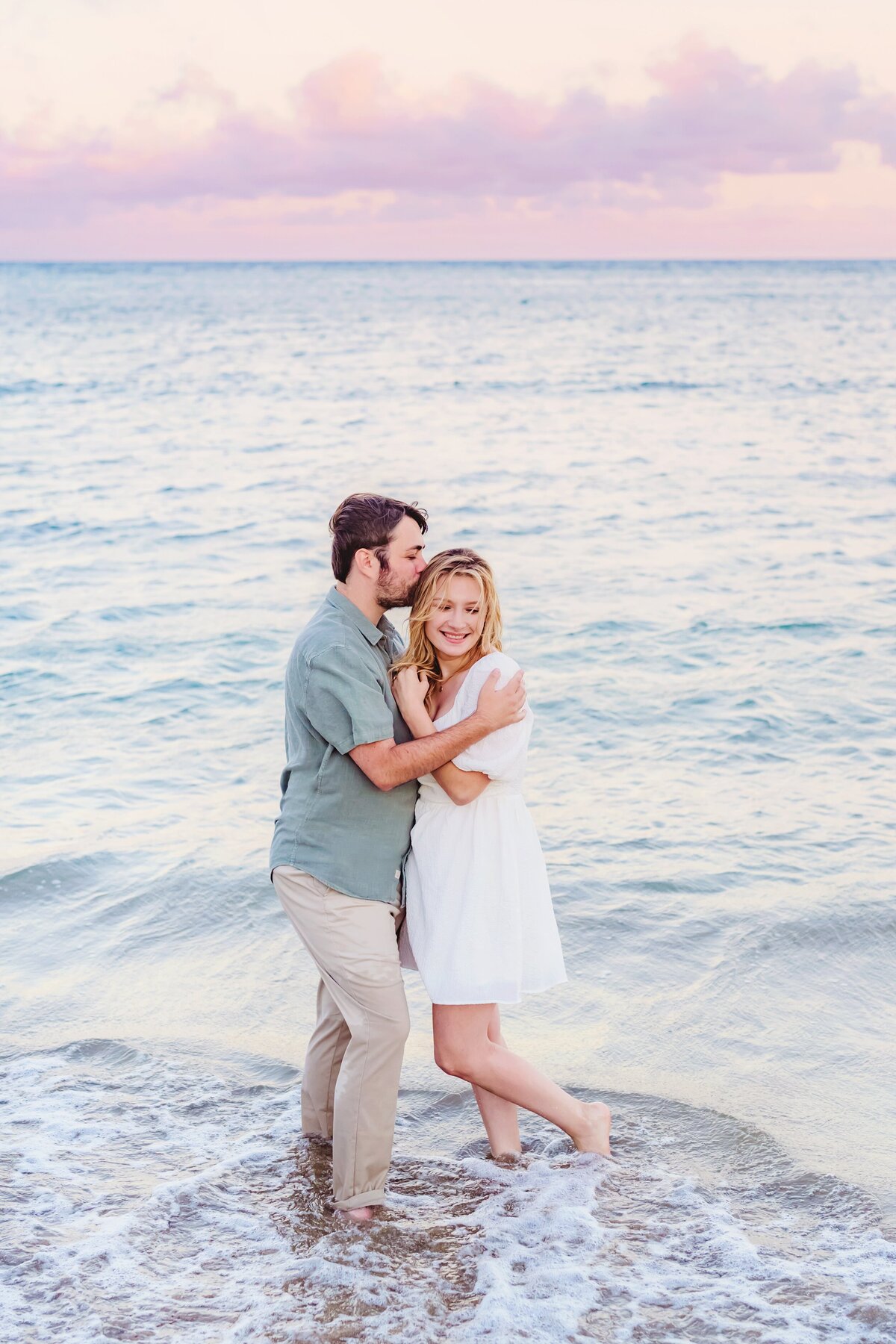 Blonde woman in a white dress is kissed by her boyfriend on the head as they stand ankle deep in the water during their Oahu couples photoshoot