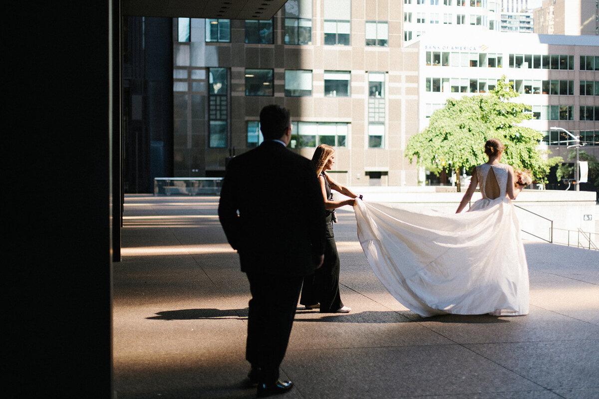 A bride in a white dress is captured by a Toronto wedding photographer as she walks outdoors in an urban setting, accompanied by two people. One of them holds her dress train while sunlight casts graceful shadows on the pavement.