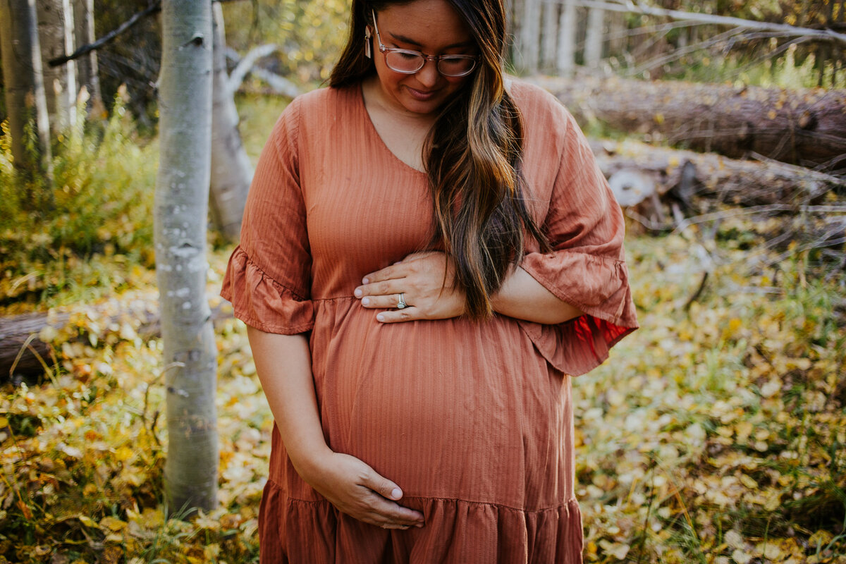 Pregnant mama holds baby bump while standing in grove of golden aspens.