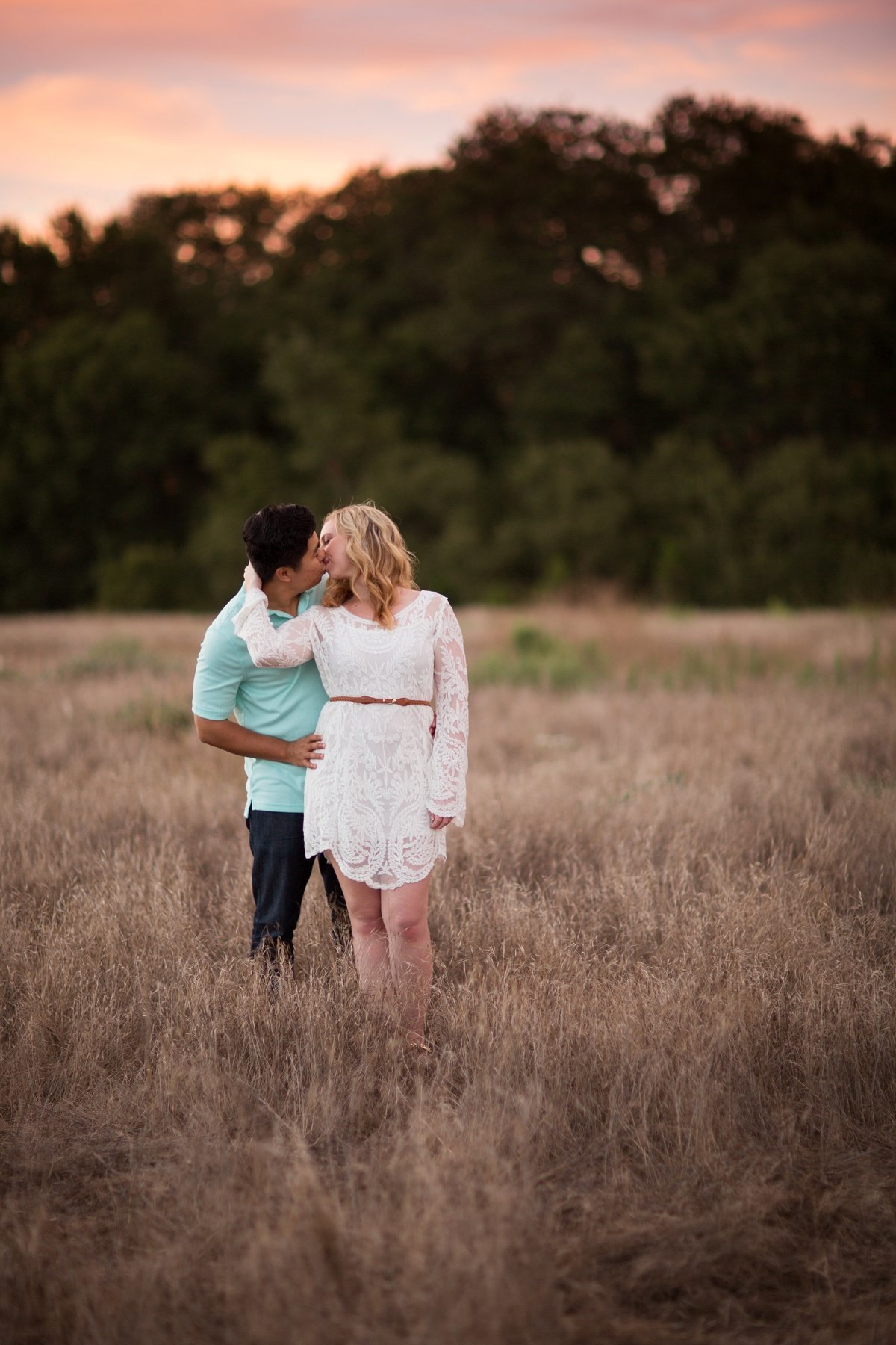 Groom to be looks around his Bride and they share a kiss as he holds her hip during photo session in an open field of brush