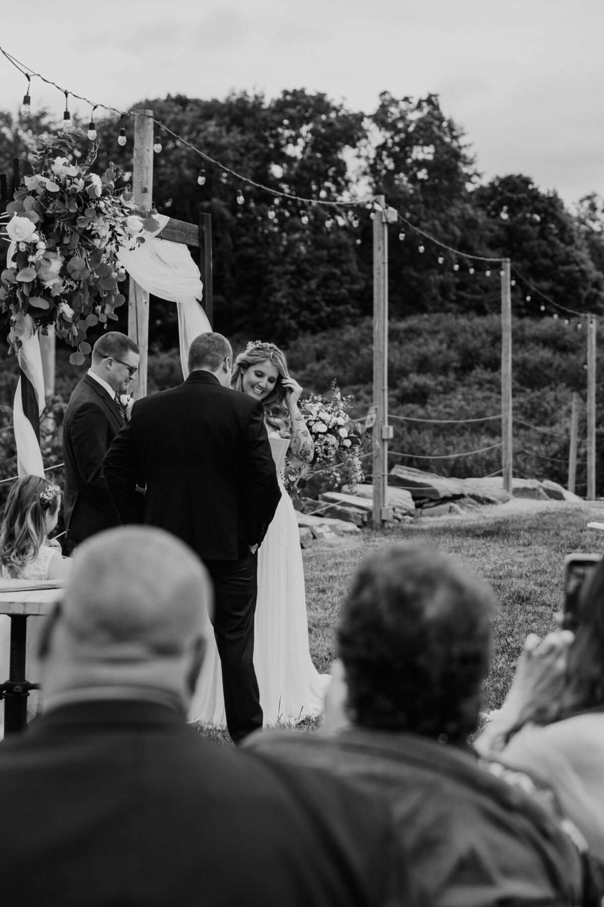 A Hudson Valley wedding photographer captures a tender moment at Locust Grove Brewery with a bride and groom exchanging vows under a wooden pergola, adorned with flowers and string lights.