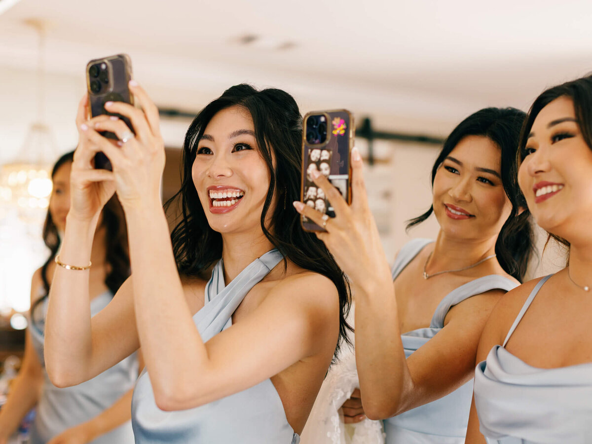 Three women in matching light blue dresses smile and capture photos with smartphones, radiating joy and excitement in a well-lit indoor setting.