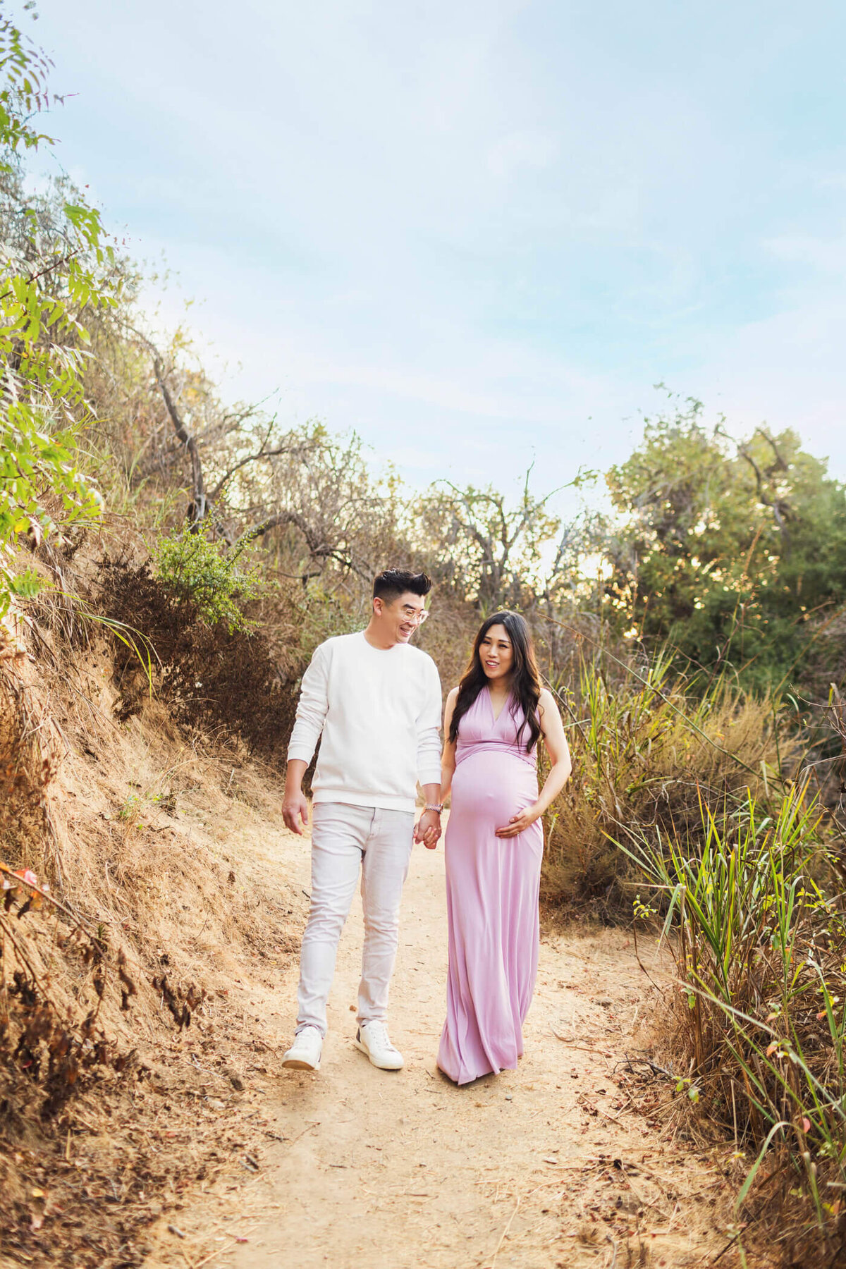 A pregnant woman walks hand-in-hand with her partner along a dirt path in a natural outdoor setting. She is wearing a form-fitting, sleeveless lavender dress that highlights her baby bump, while he is dressed in a white long-sleeve shirt and light pants. They both smile and look at each other affectionately. The surrounding greenery and blue sky create a serene and joyful atmosphere.