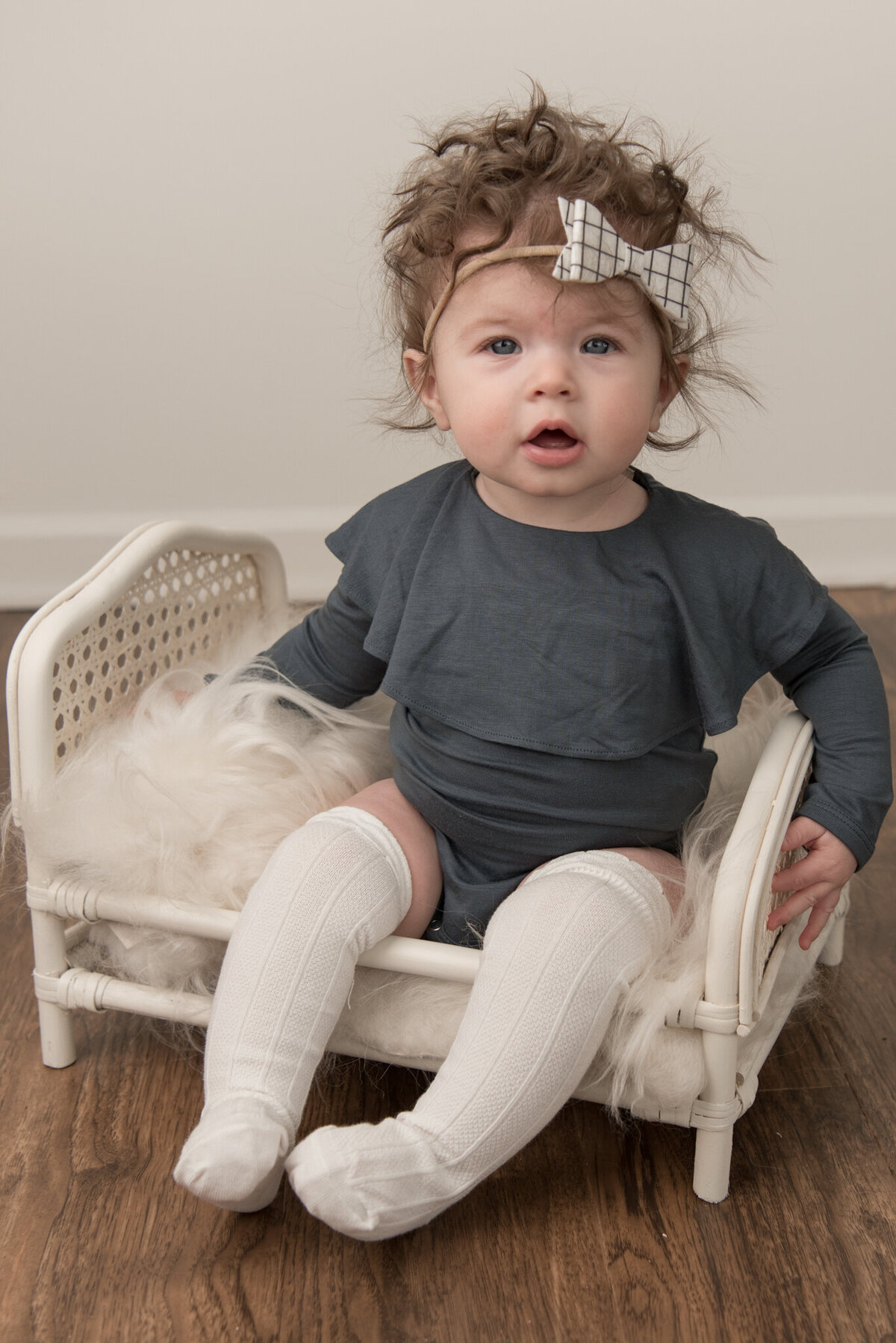 Six month old girl sitting on white bed with white headband and gray romper