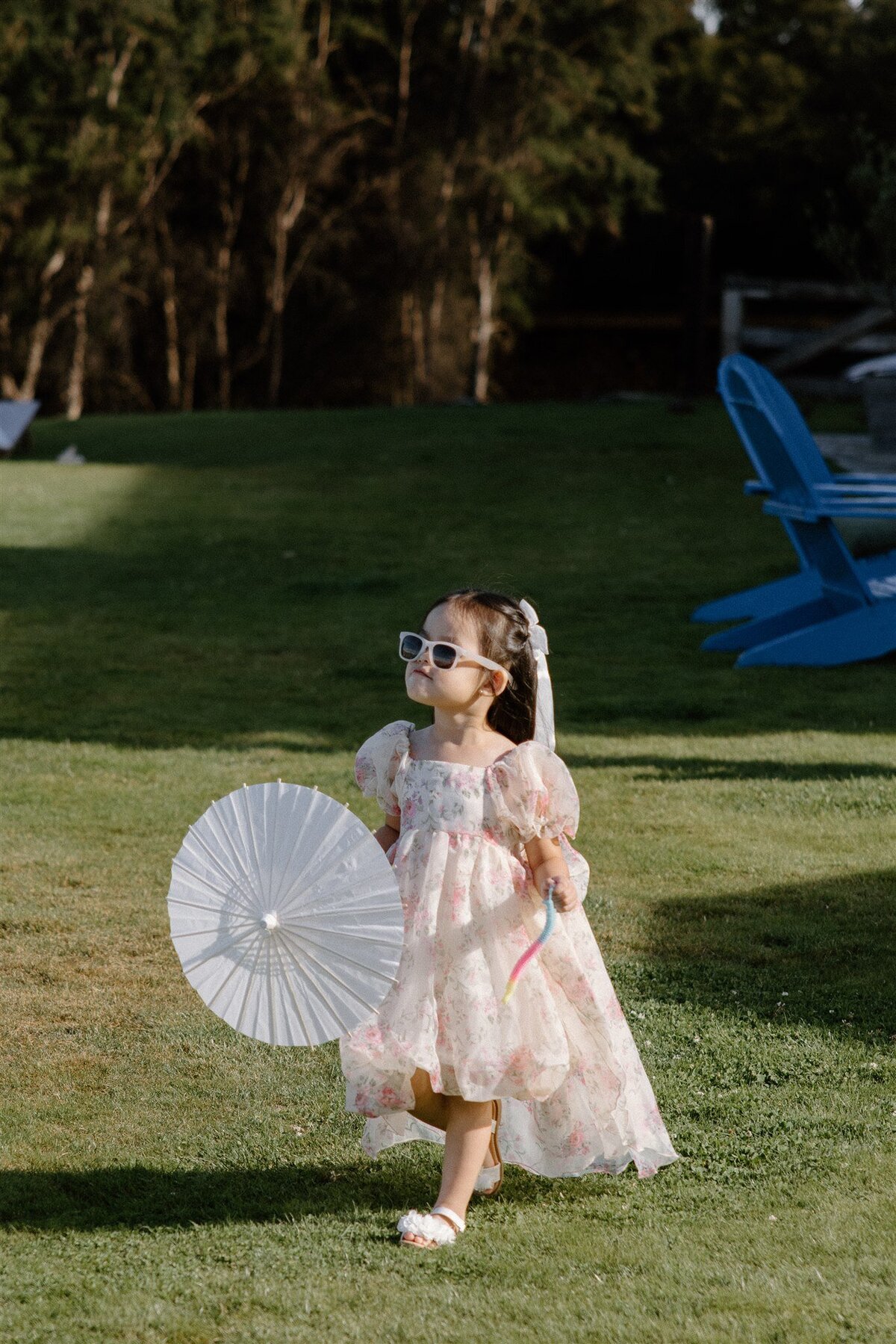 Young flower girl walking across a Wanaka lawn in a pink floral dress and sunglasses, holding a white paper parasol at a summer alfresco wedding.