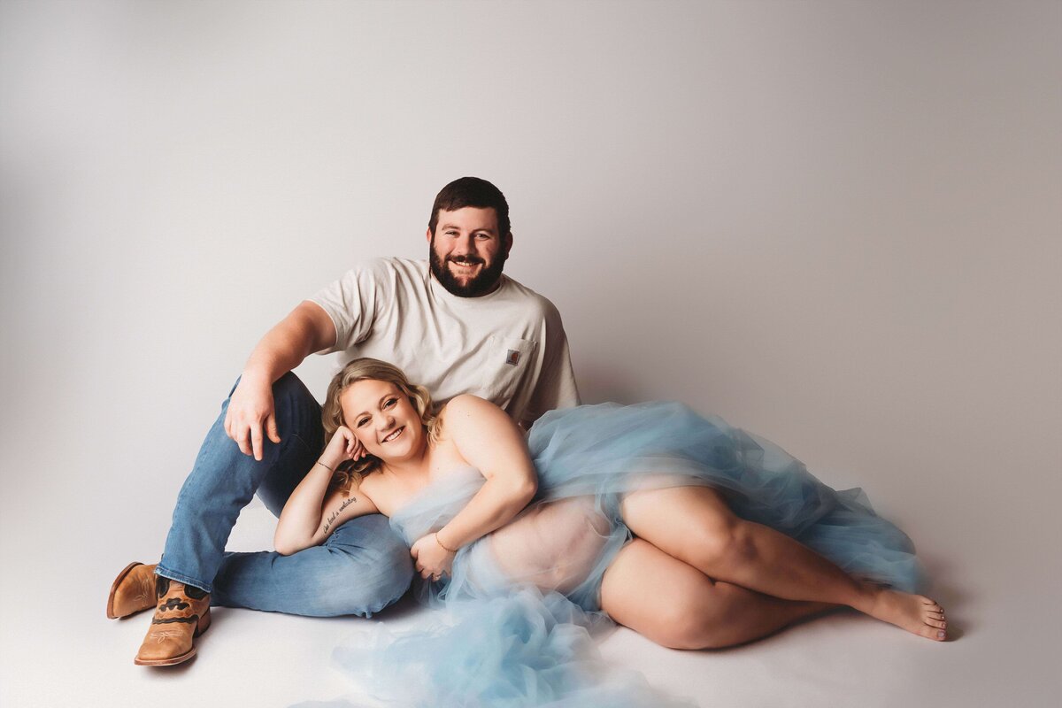 Maternity photo of a smiling couple, with the woman wearing blue tulle , leaning on her husband's leg who is wearing boots, blue jeans, and a t shirt.
