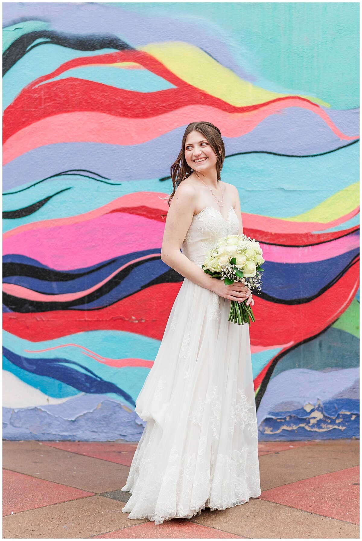 Bride stands with her body facing right and her head looking off to the left while holding her bouquet in her arms while standing in front of a colorful wall in downtown Denver outside of the venue Skylight