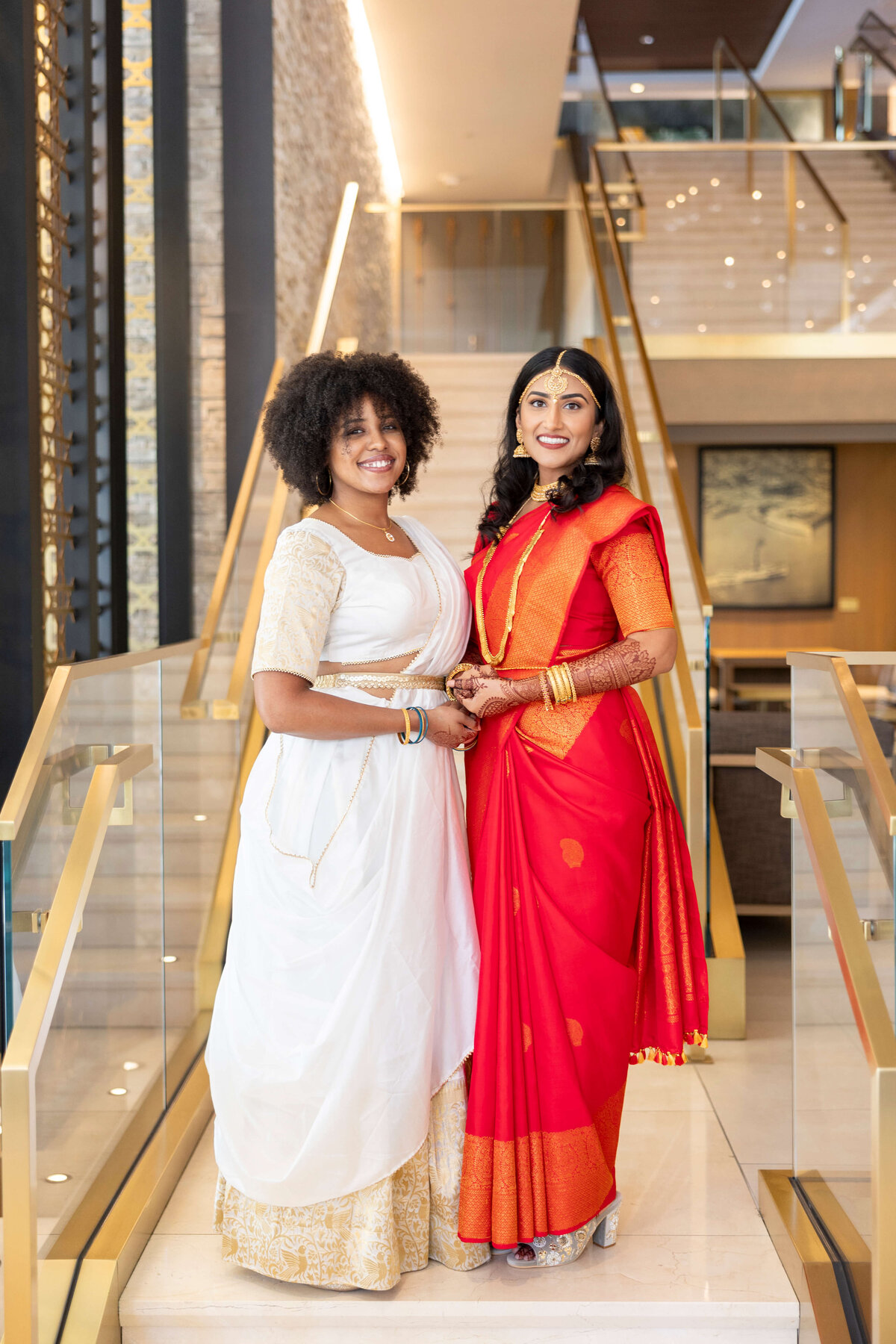 Two women stand on a staircase, smiling. One wears a white saree with gold details, and the other wears a red saree with gold trim. The background features a modern, elegant interior with a stone wall and glass railing.