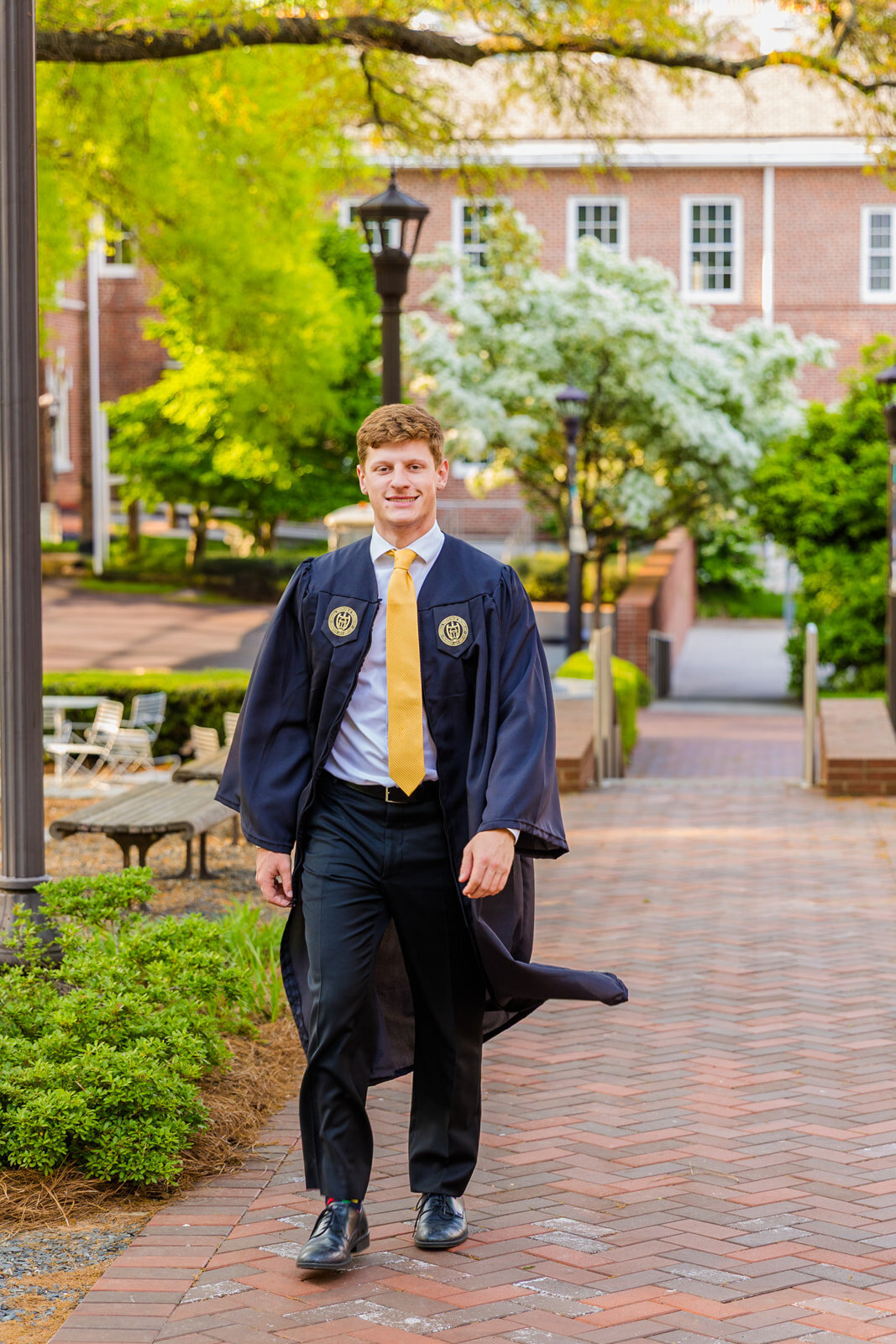 Senior walking on Georgia Tech campus in front of building and blooming trees wearing graduation gown over a white shirt and a yellow tie during graduation photo session with Atlanta photographer Laure Photography