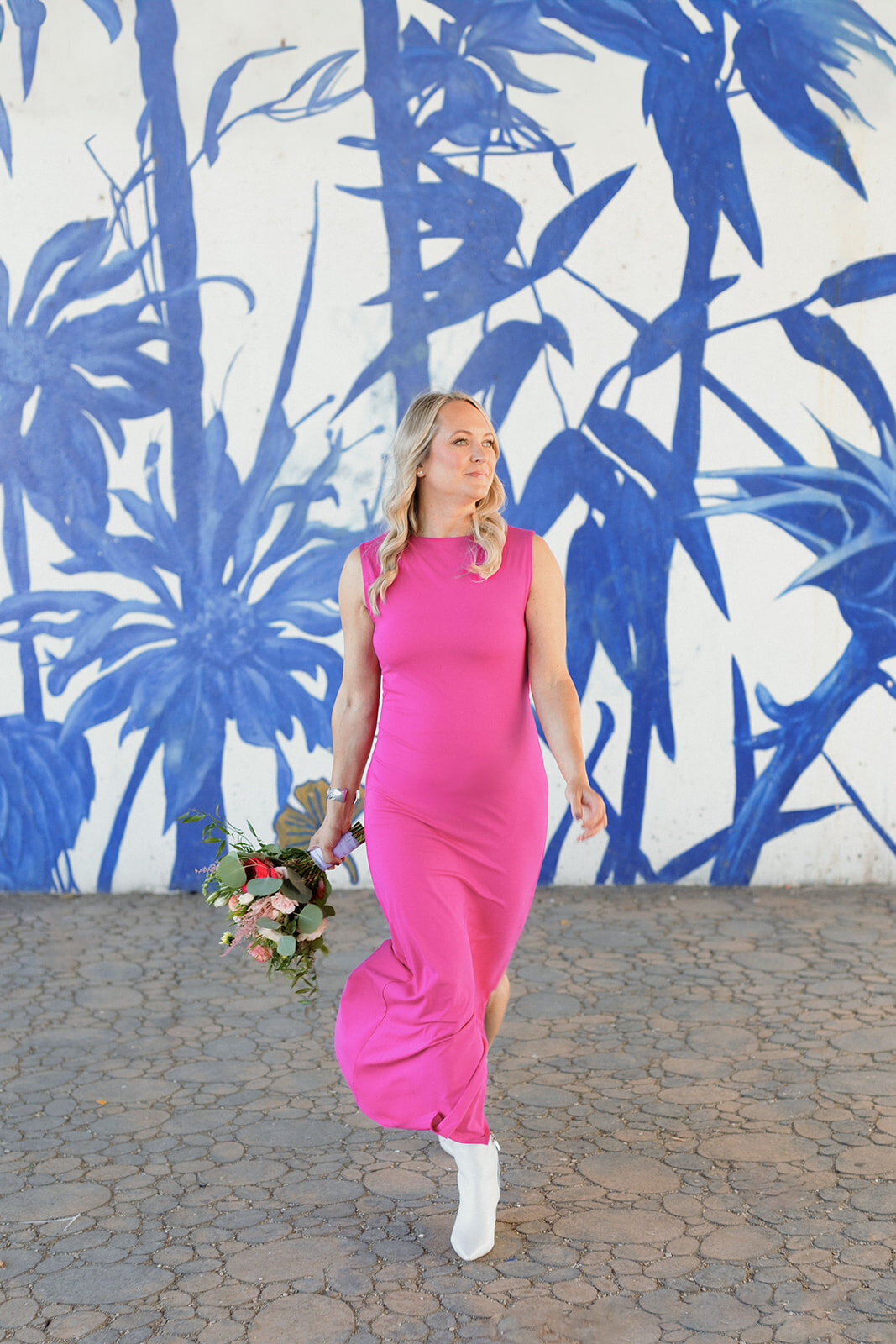 blonde woman walks in fitted pink dress toward the camera in front of a blue floral mural. She holds a pink and red bouquet and looks off to the side.