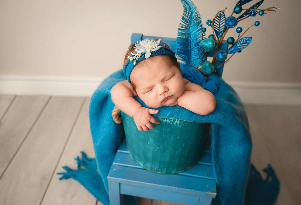 Infant girl posed in a blue scene in a bucket sitting on a chair prop and accessories.