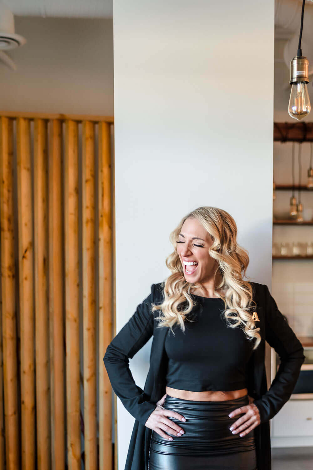 Woman laughing and leaning against white wall in black skirt and top at local Kelowna yoga studio.