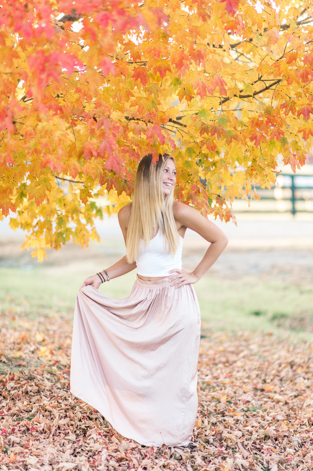 Young woman holds her dress as she smiles under a colorful tree during fall.