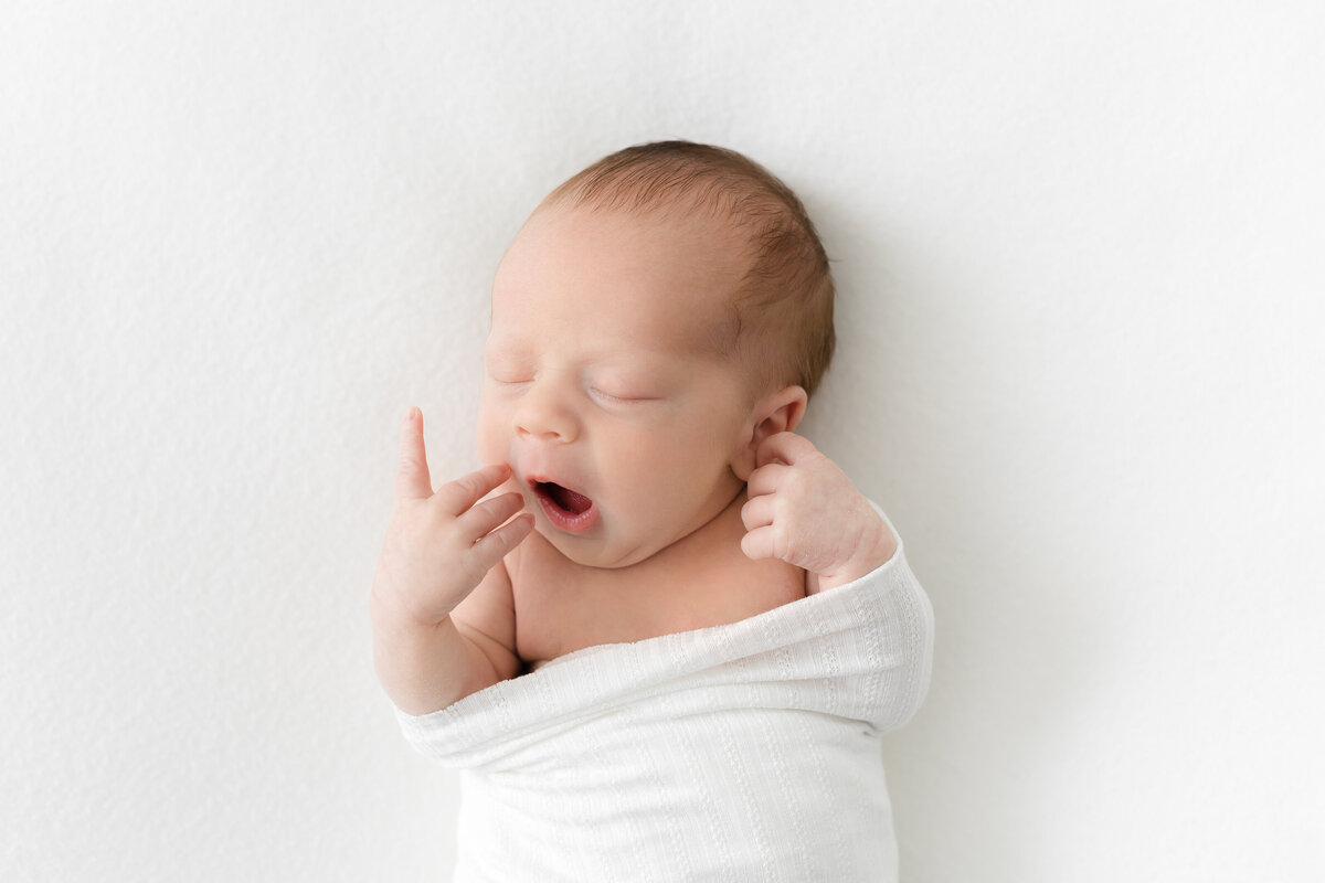 A dc newborn photography photo of a sweet baby boy swaddled up in a white blanket yawning and stretching