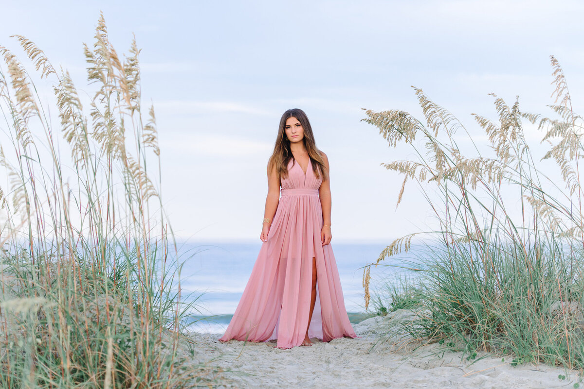High school senior girl in pink dress standing in beach grass on Sullivan’s Island at sunset