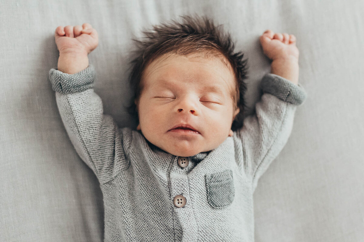 An adorable newborn baby boy with a full head of hair lays on a bed with his arms stretched straight into the air