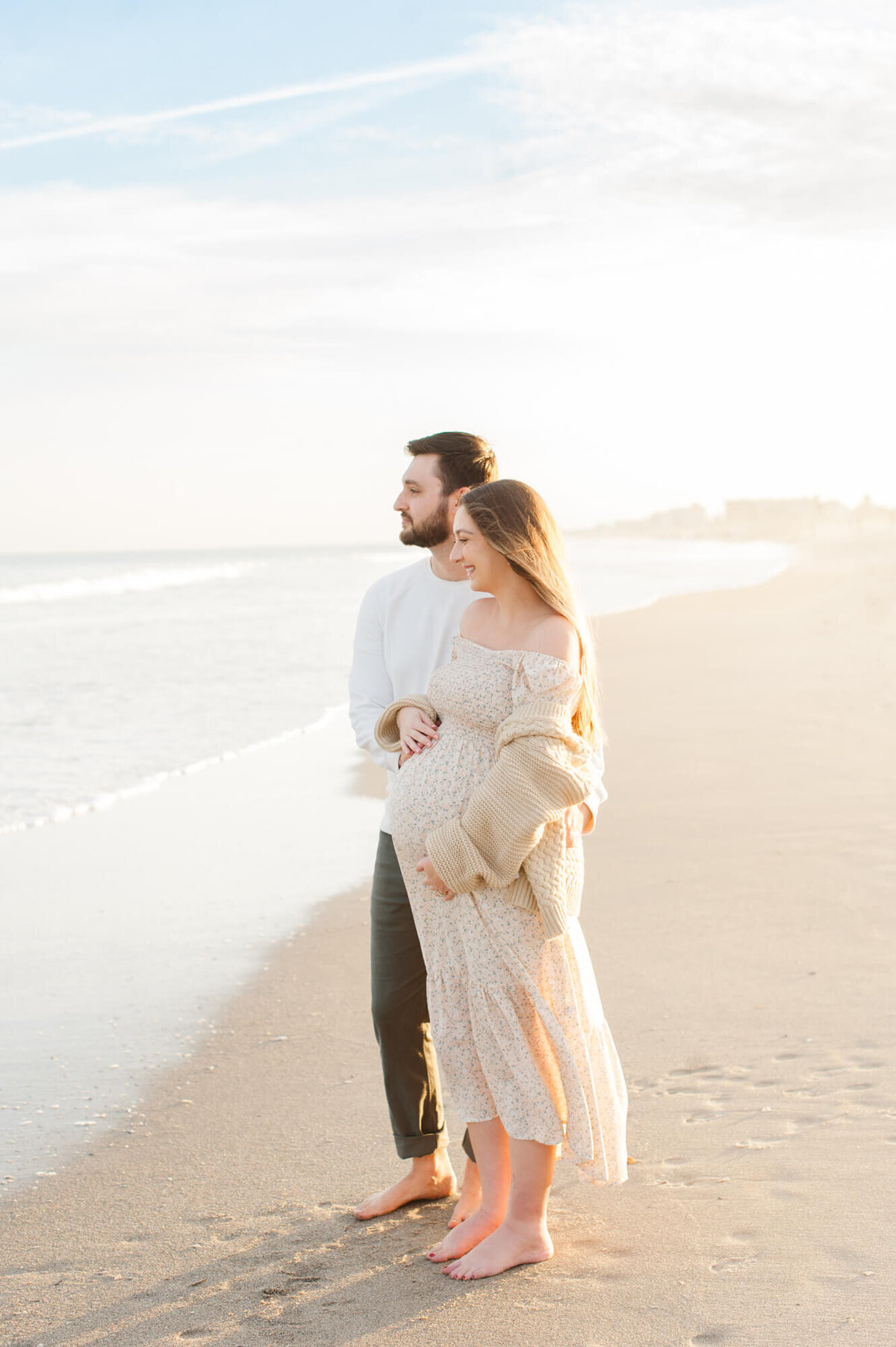 New parents holding moms belly and looking out at the ocean during her orlando maternity photographer session