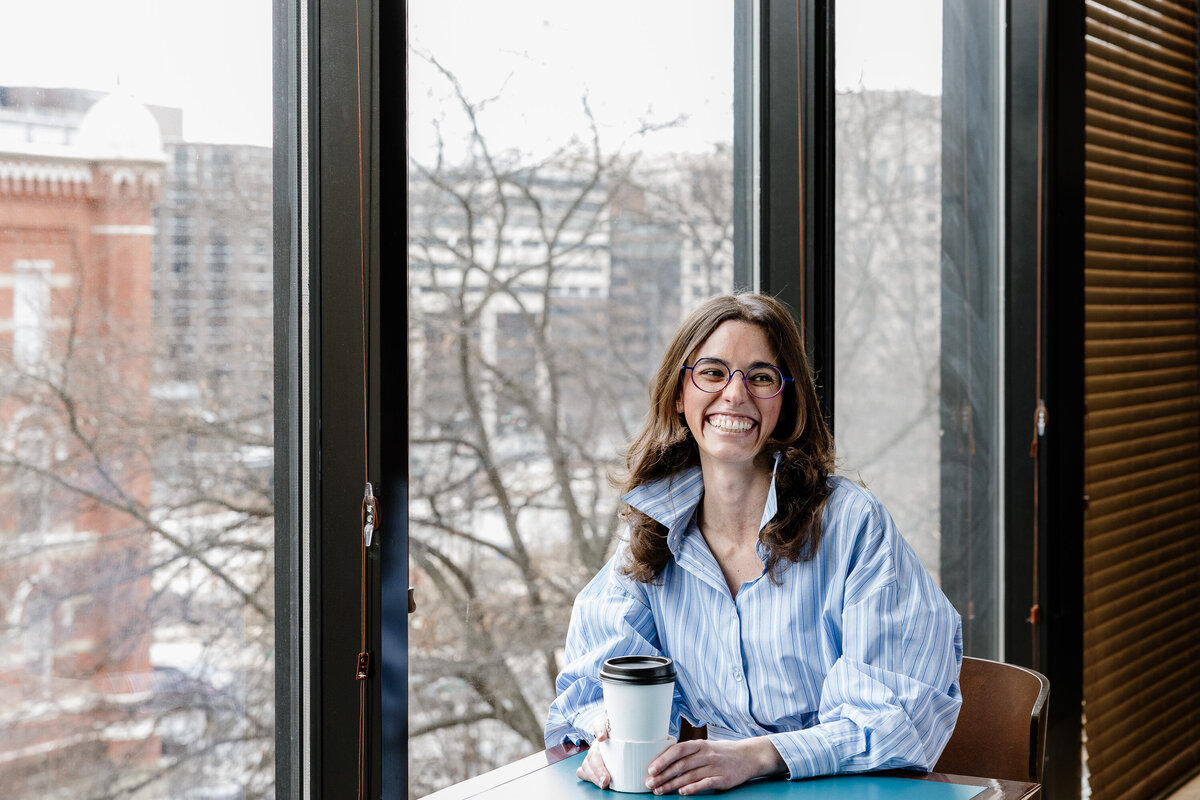 A young woman sitting in front of a window with a cup of coffee, laughing and smiling. posed but also candid 