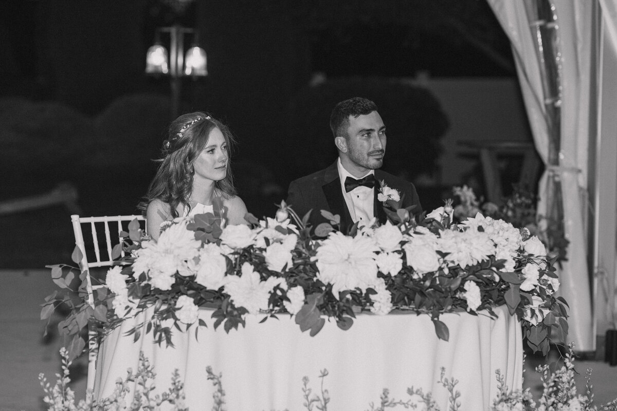 Bride and groom sit at a beautifully decorated sweetheart table during their wedding reception, surrounded by lush floral arrangements. The bride, wearing a delicate headpiece, and the groom, in a black tuxedo, share a thoughtful moment as they listen to speeches. The black and white photo captures the elegance and intimacy of the evening.