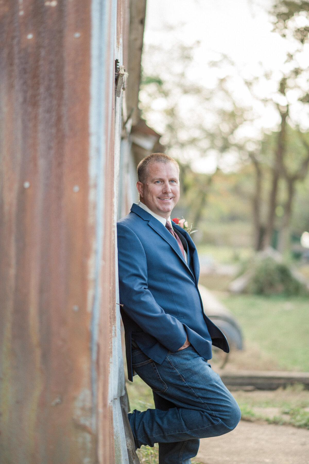 The groom stands with his foot propped up on the side of a rusty metal barn, smiling at the camera