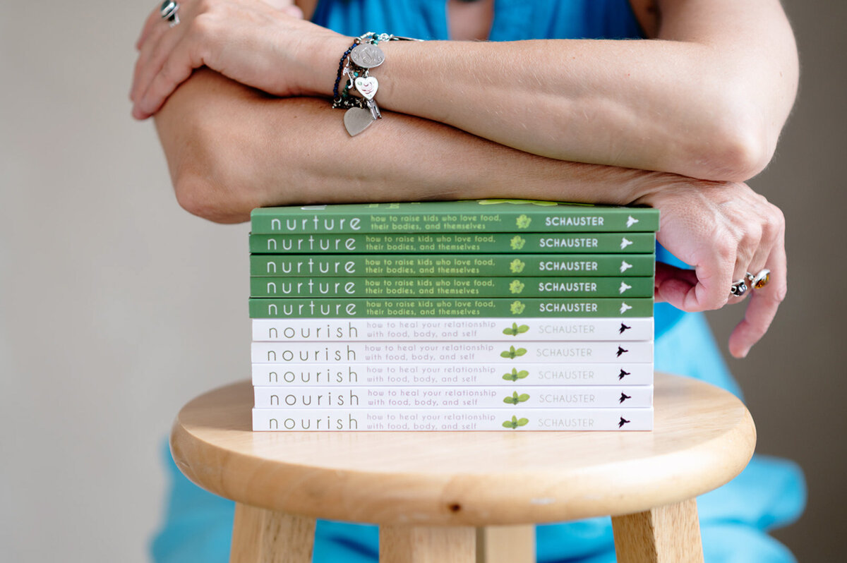 A person in a blue dress rests their arms on a stack of books titled "Nurture" and "Nourish" by Schauster, seated on a wooden stool, conveying calm and focus.