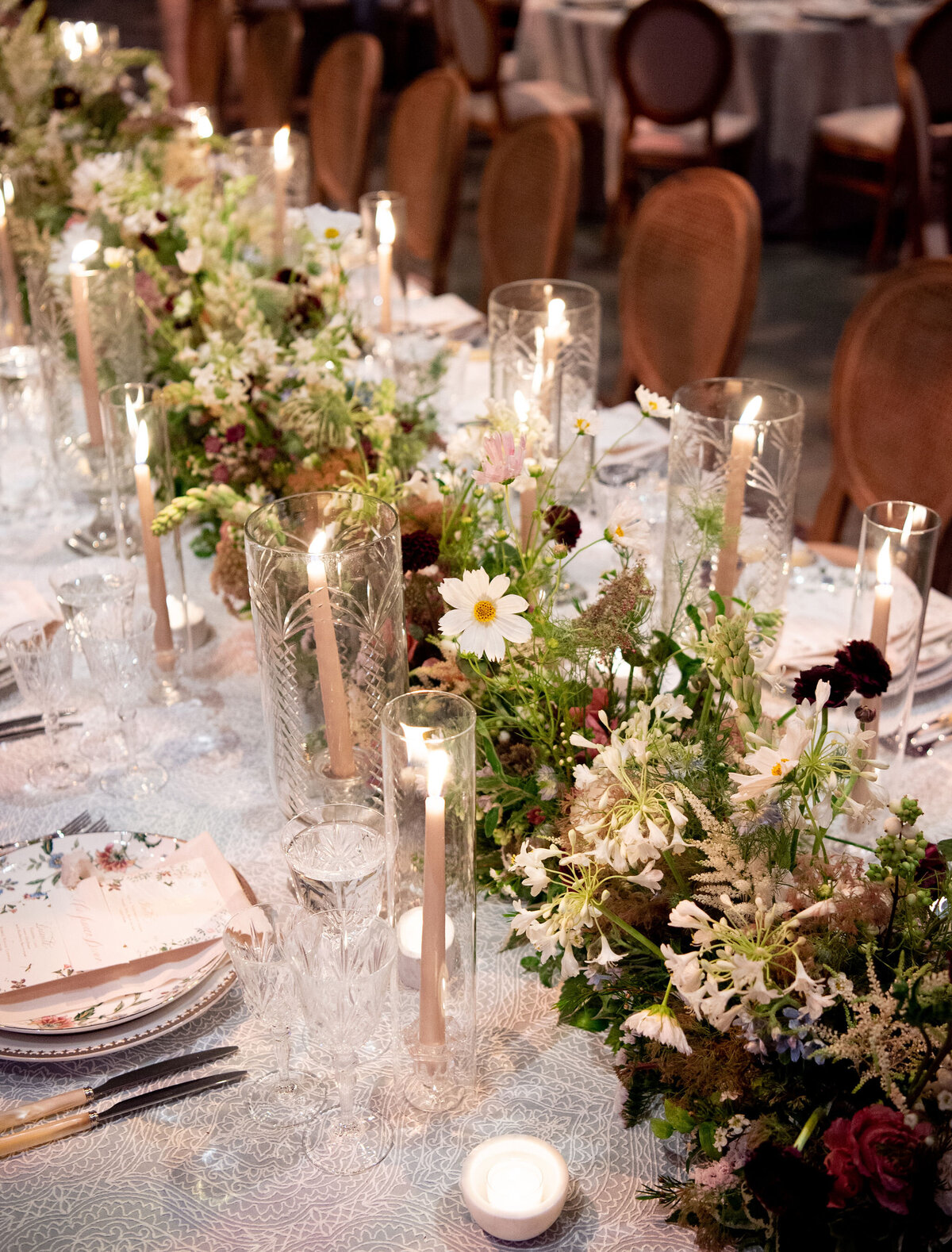 This exquisite setting was captured at Washington National Cathedral in Washington D.C. by talented photographer Twah Dougherty and filmed by Current Films as Videographers. The stunning image showcases a beautifully set dining table adorned with floral arrangements and lit candles in tall glass holders, adding to the elegant and romantic atmosphere. The tableware features patterned plates, crystal glasses, and neatly placed silverware, enhancing the overall charm of the scene.