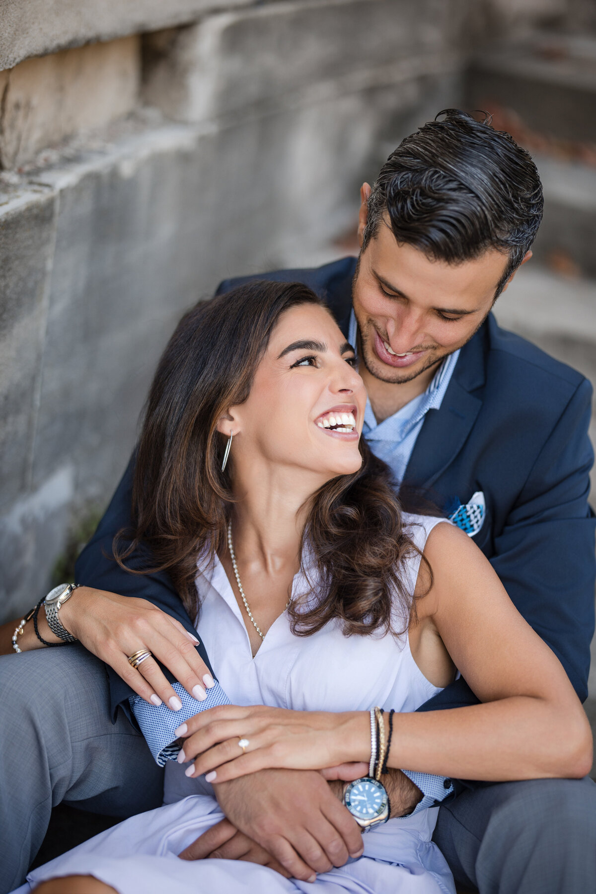 A couple sitting closely, smiling at each other warmly. They are wearing casual formal attire at their Untermeyer Gardens engagement photo shoot