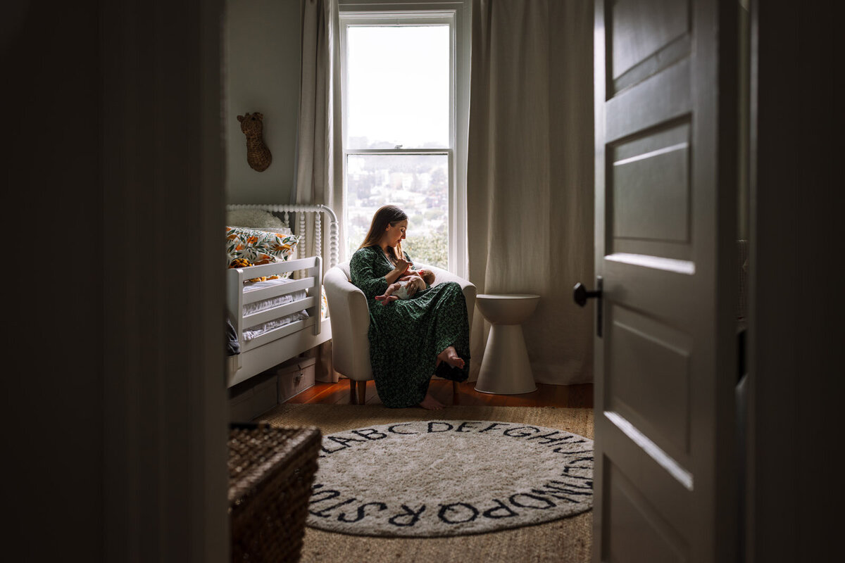 mom in green dress sitting in chair in nursery in front of window breastfeeding her newborn