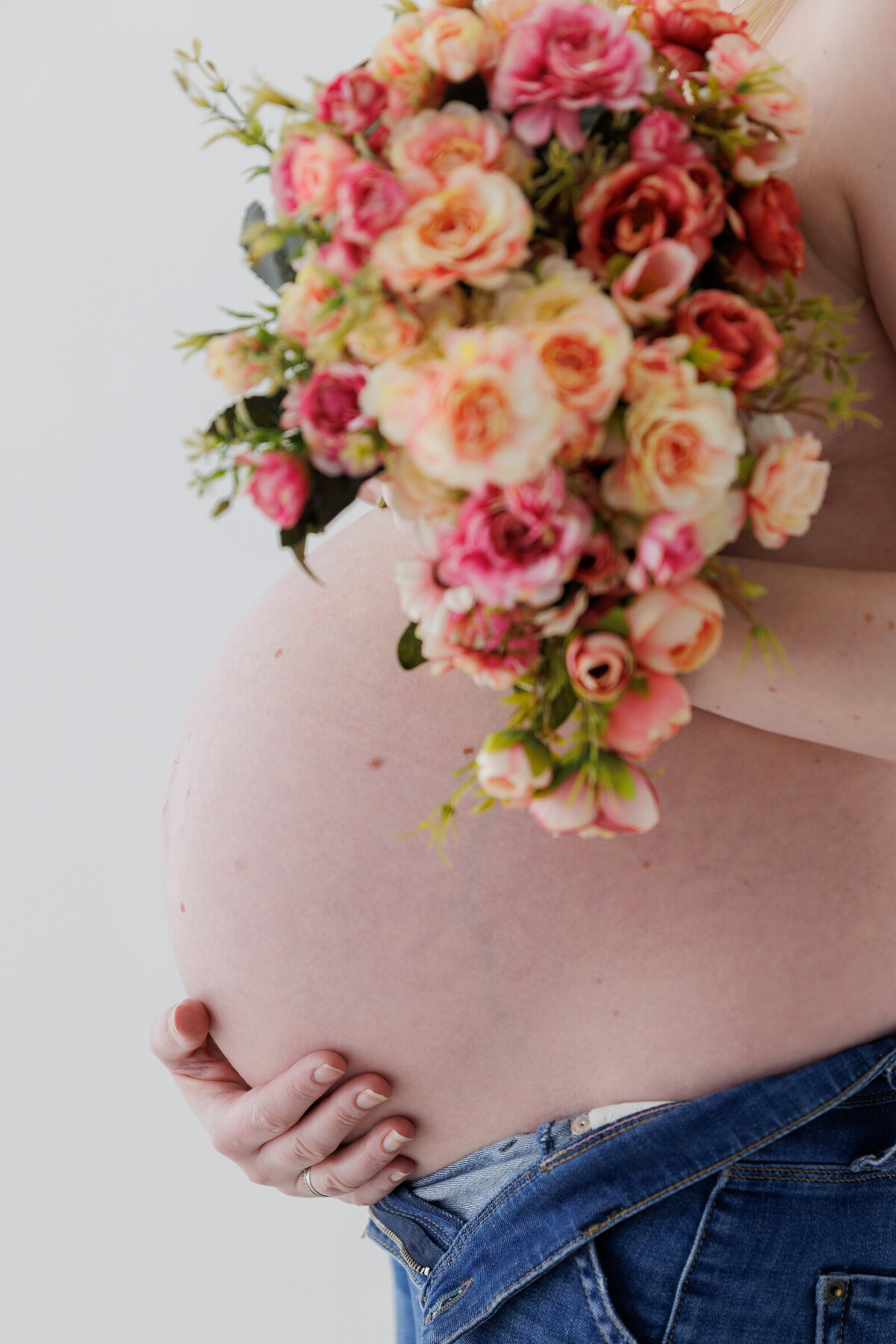 Gravidmage fotografert i profil, en blomsterbukett holdes over brystene og man kan se dongeribuksen er åpen for å gi plass til magen. Fotografert i Studio Landmark, Fyllingsdalen.