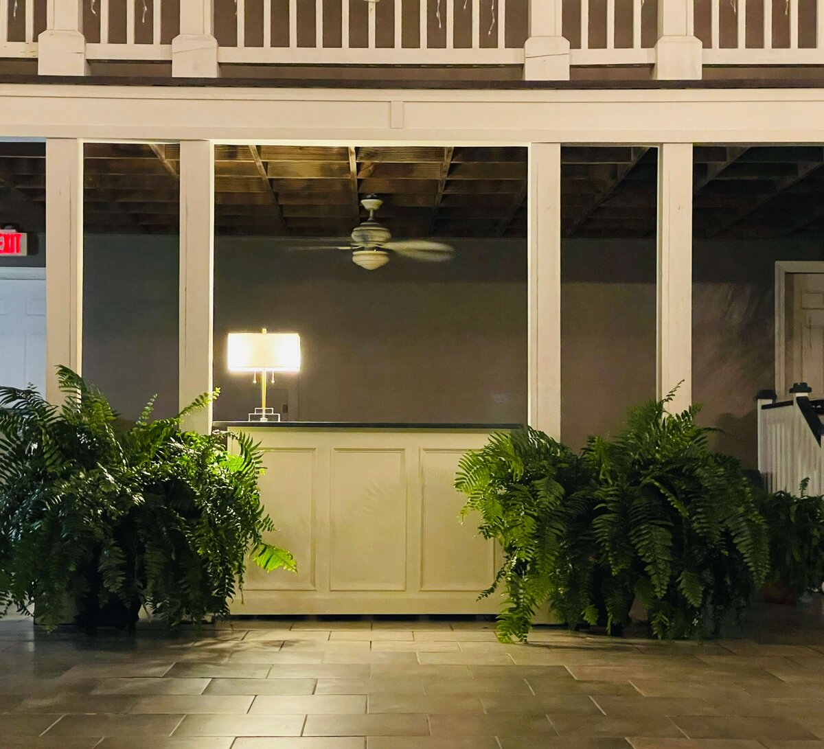 A new bar table with a lamp stands under a balcony with lush green ferns in a courtyard.
