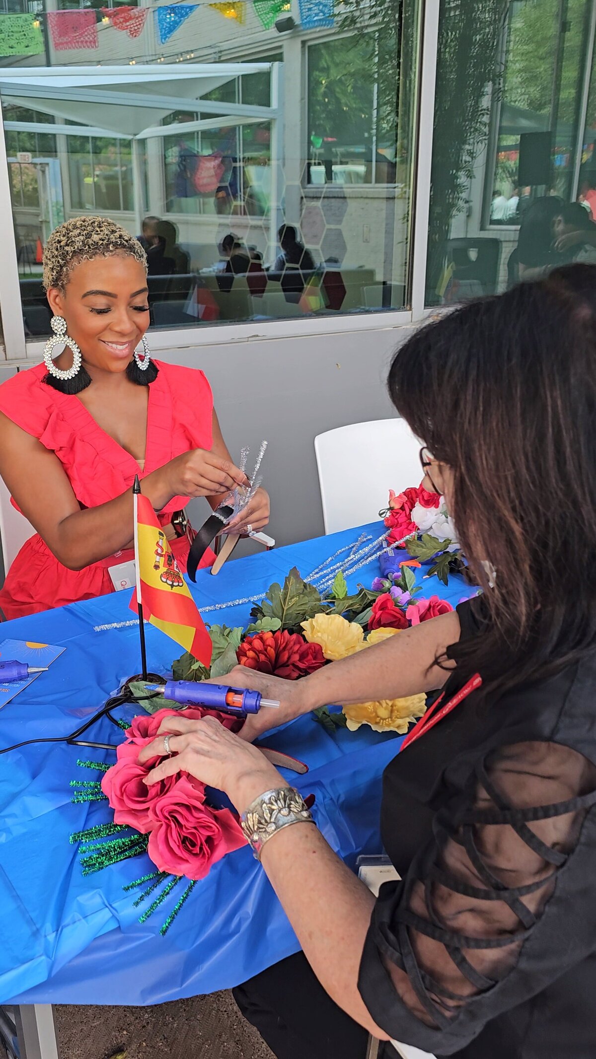 The world of craftsmanship as a woman in a stunning red dress delicately glues vibrant flowers onto a wreath in our artistic workshop. The long table is adorned with a spectrum of materials, and in the foreground, witness the beauty of the final piece—a masterpiece of colors and creativity.
