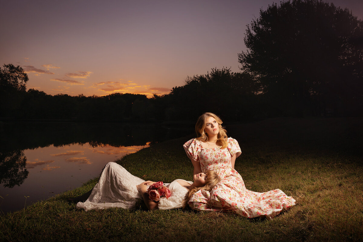 Two senior girls in floral dresses pose by a lake at sunset, captured by a Dallas senior photographer.