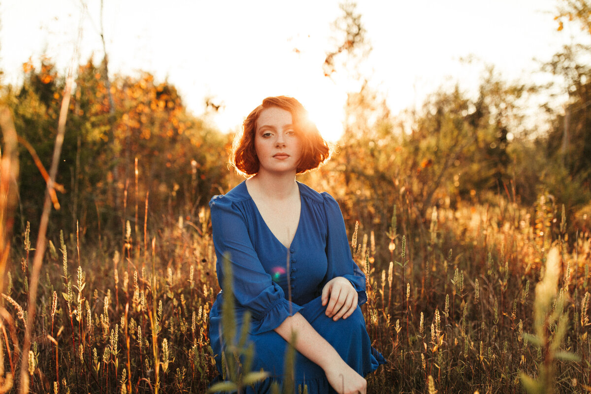 High school senior in long blue dress squatting in a golden field at sunset