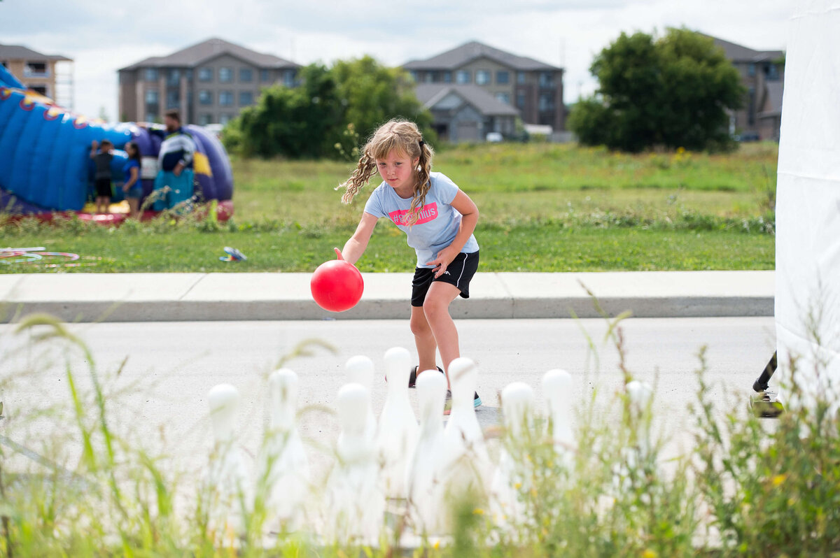 a little girl throwing the red bowling pall to knock down pins as part of a children's activity at a corporate children's event.  Captured by Ottawa Event Photographer JEMMAN Photography COMMERCIAL