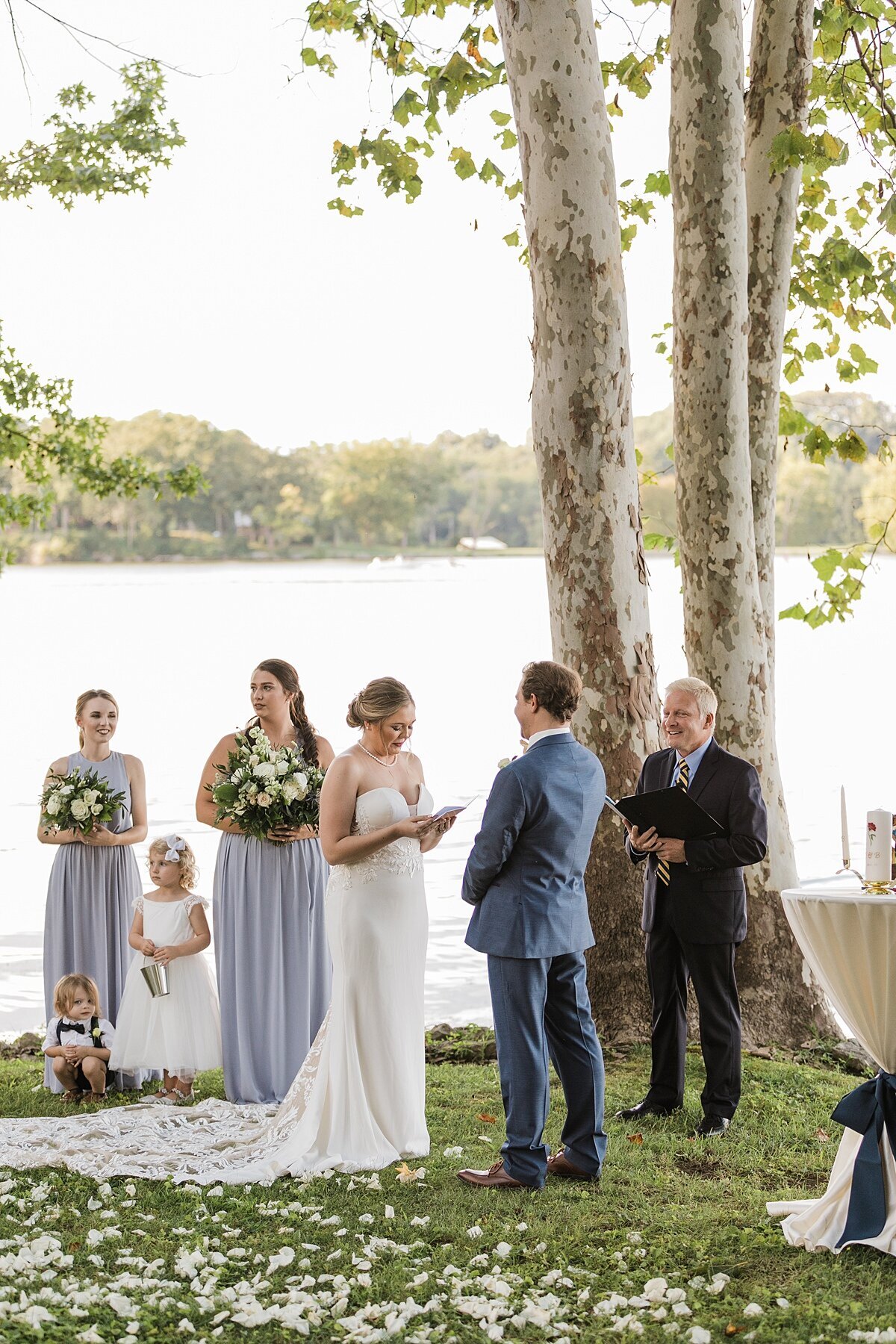 Bride wearing a strapless lace wedding dress reads her vows to the groom wearing a grey suit as they stand lakeside at The Estate at Cherokee Dock. The bridal party, dressed in light blue holding bouquets of white and yellow flowers stand on the lawn covered in white flower petals.