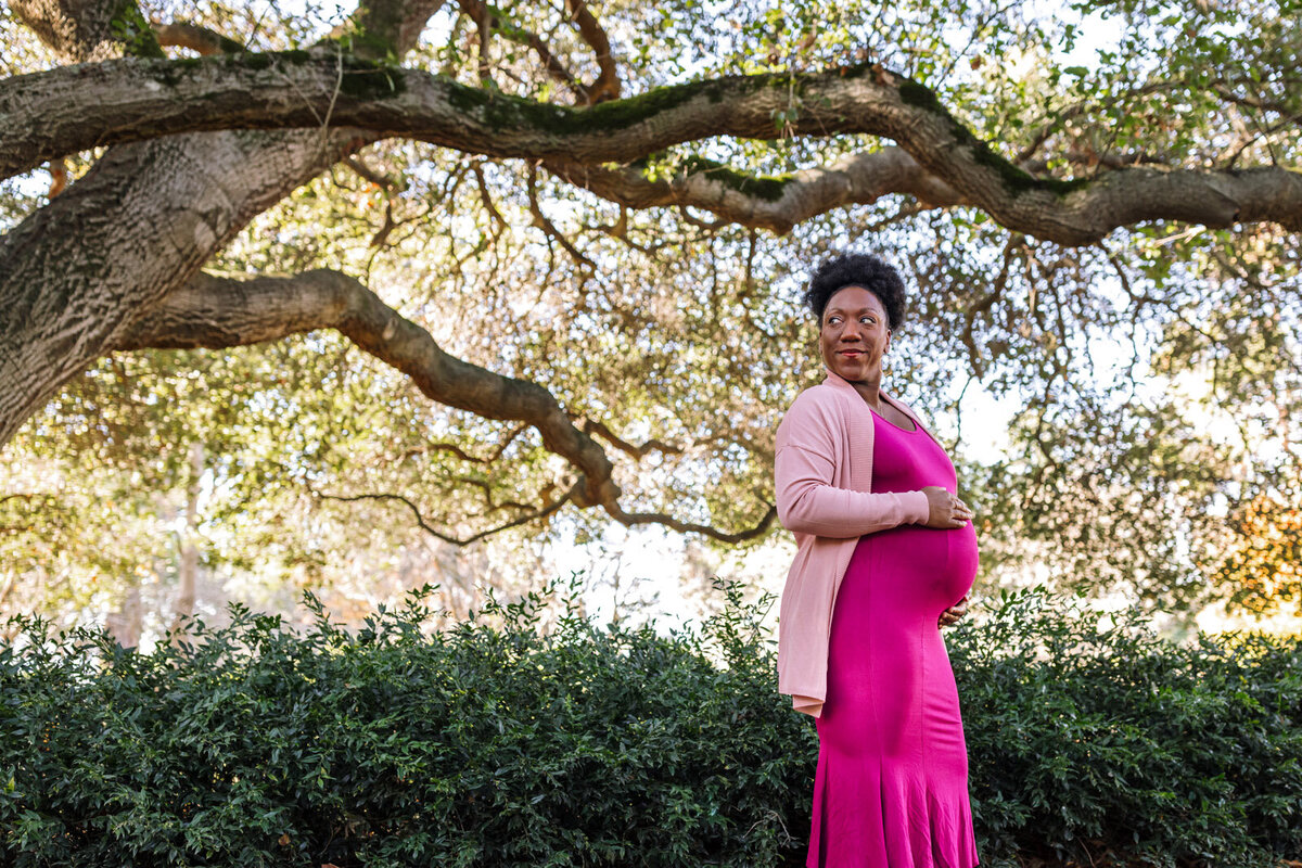 pregnant woman in hot pink dress standing under tree branches in front of greenery smiling and looking over her shoulder