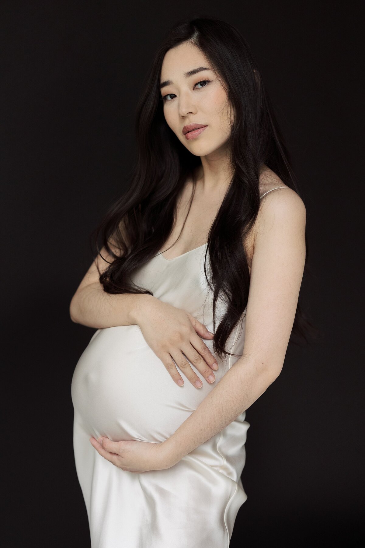 Pregnant woman holds belly posing in cream silk dress.