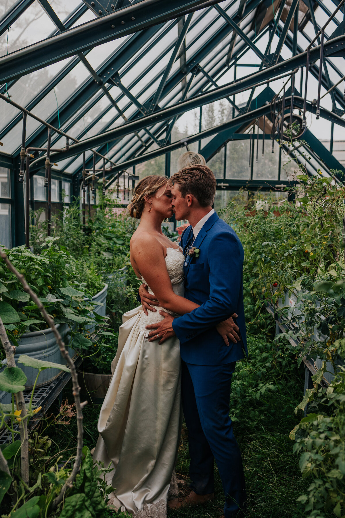 Bride and groom kiss each other in the middle of full grown greenhouse.