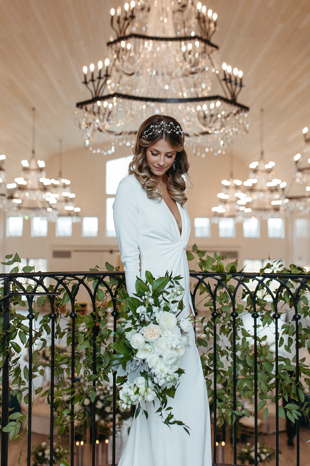 Bride in a white wedding gown stands in front of rail wrapped in garland holding a white bouquet with a chandelier above.