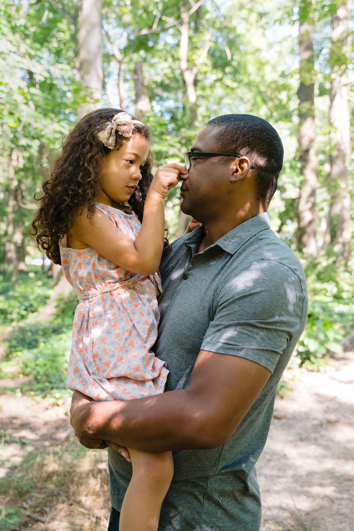 A person holding a small child in their arms as she pinches their nose while they stand in a wooded area.
