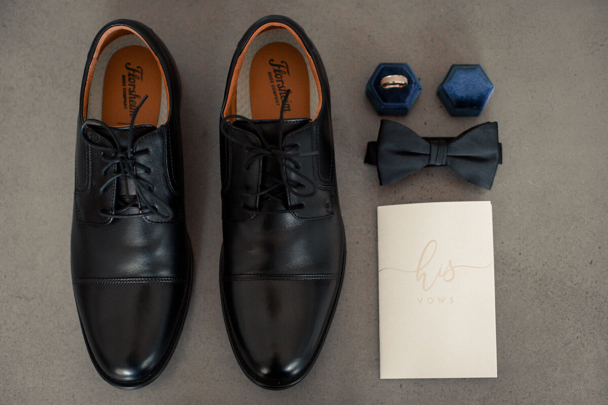Groom's black dress shoes, bow tie, and wedding details arranged on a grey surface