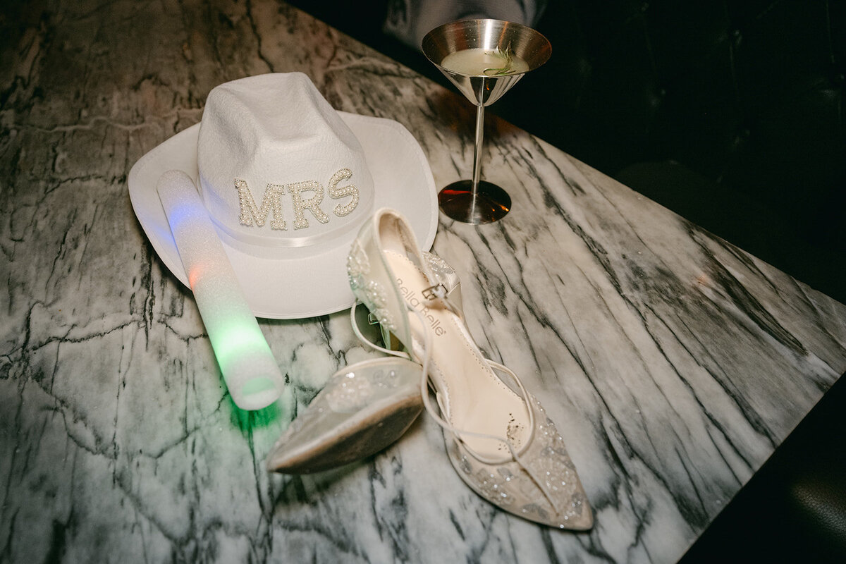 Bridal detail flat lay featuring custom "Mrs" hat, glittery Jimmy Choo heels, glow stick, and martini glass on luxury marble bar
