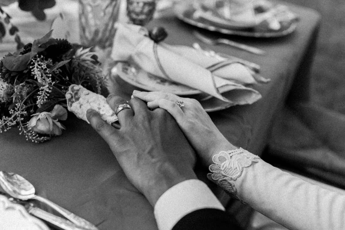 Black and white photo of a couple holding hands at their wedding reception, surrounded by elegant table settings. The image captures the intimacy and details of the reception, including the delicate floral arrangements.