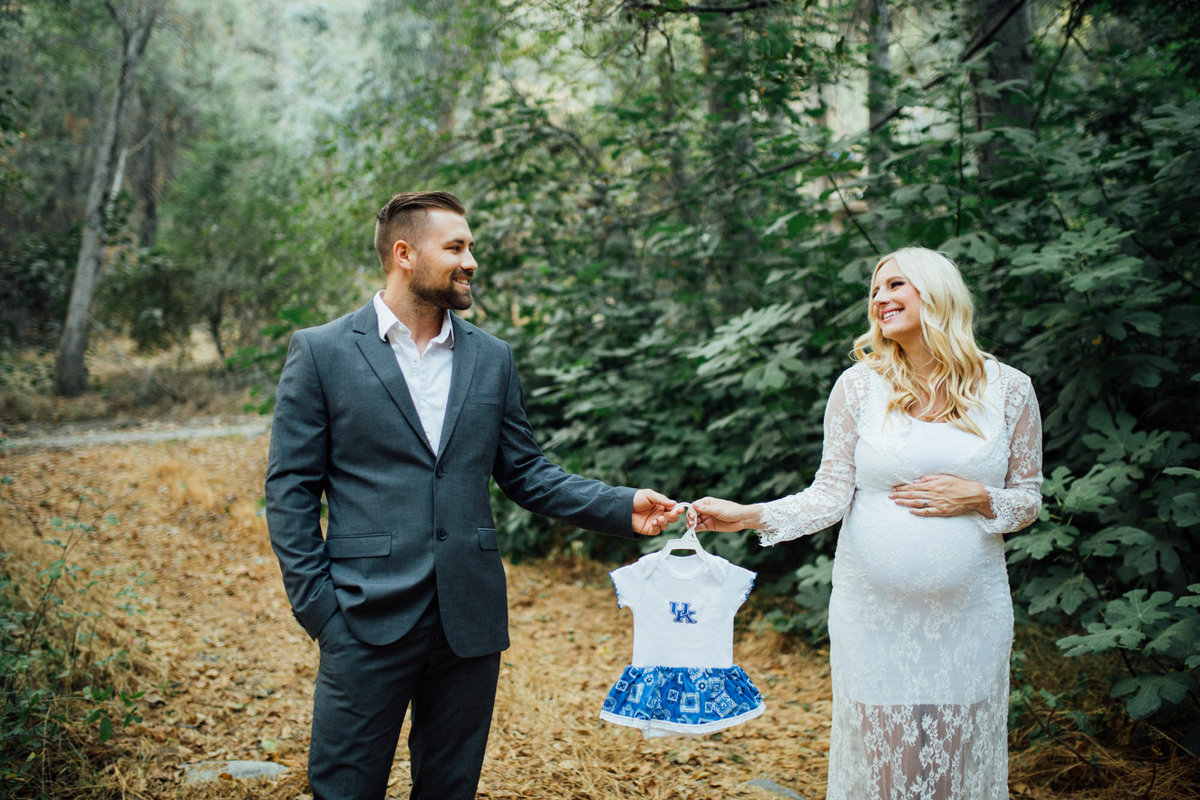 Parents to be stand in the mountains looking at each other while holding a little dress for their soon to be born daughter