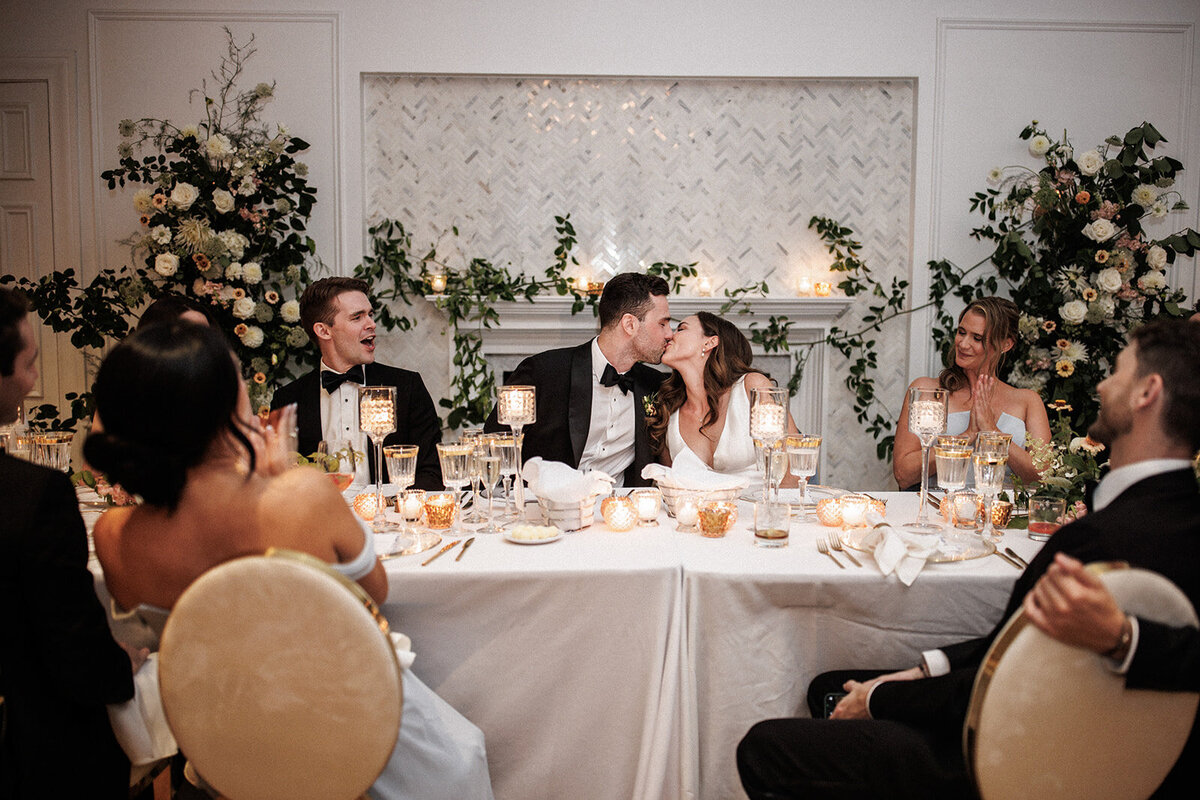 A couple kissing at their wedding reception at the Atwater Club, framed by garden flower arrangements.