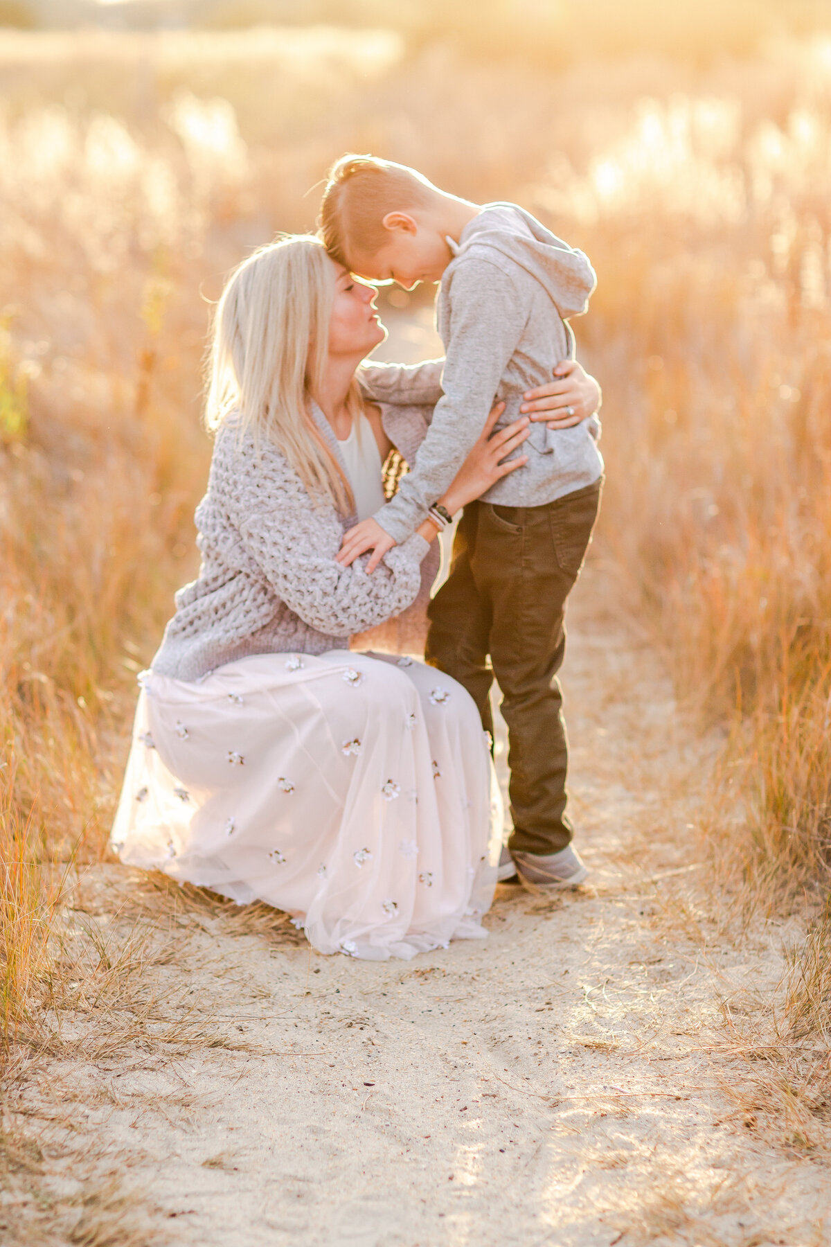 A mother kneels in a sunlit field as her young son leans in close, their foreheads gently touching in a tender moment of connect captured by Brooke Tucker photography