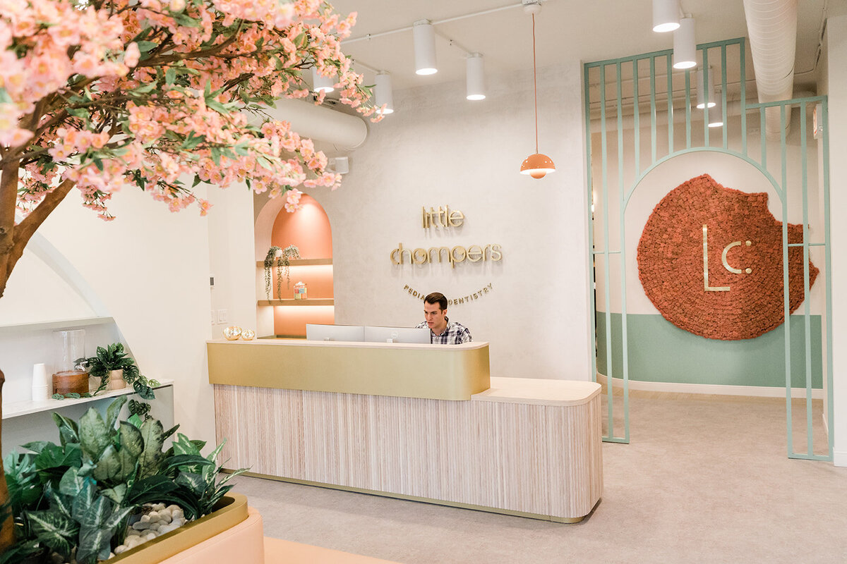 A receptionist works quietly at the Little Chompers family dentistry office in Andersonville Chicago. Cherry blossoms and a modern palette of peachy pink, muted rust, and minty sage green add an upscale element to the waiting area for this popular pediatric dentist.