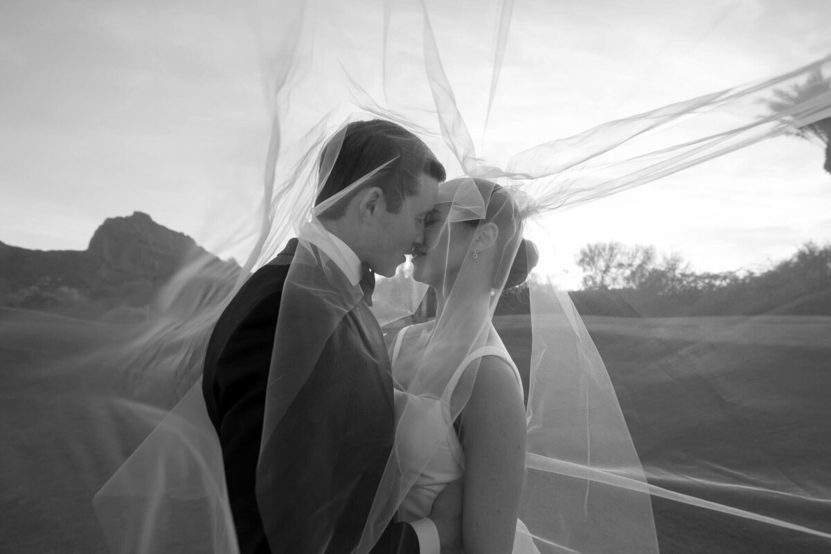 Bride and groom share a romantic kiss under the veil at sunset with Camelback Mountain behind them, captured by an Arizona wedding photographer.