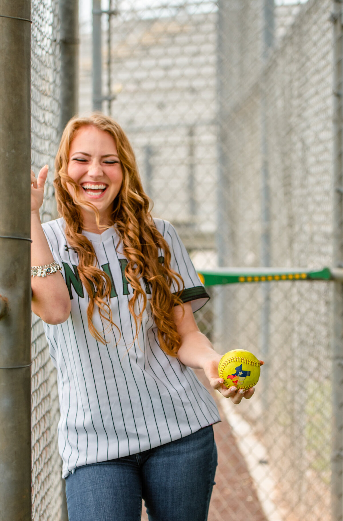 A high school softball player leans against a dugout fence and holds a softball with a big smile.