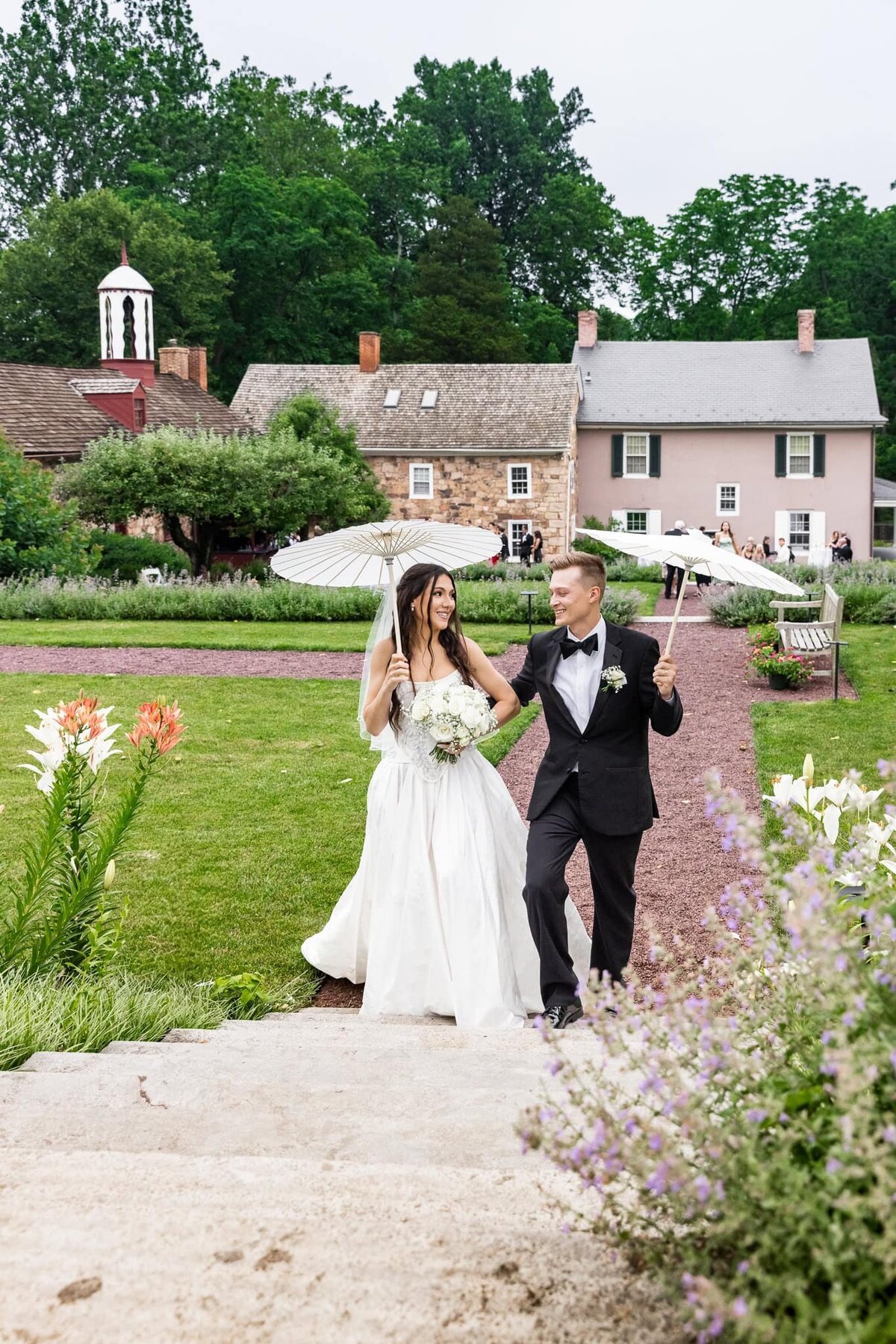 A bride and groom walk through a historic garden in Lancaster, holding white parasols and smiling joyfully at each other. The bride wears a flowing satin gown and carries a bouquet of white flowers, while the groom looks dapper in a black tuxedo. Behind them, stone and pastel-colored colonial buildings frame the lush garden setting.